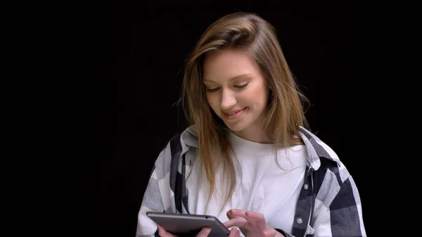 Portrait of young caucasian long-haired girl in shirt smilingly working with tablet on black background. — Stock Photo, Image