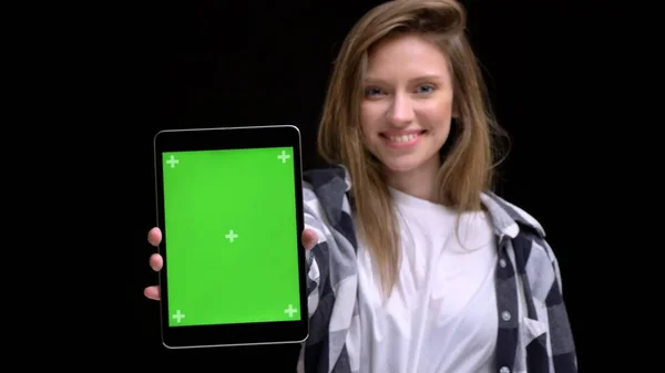 Portrait of young caucasian long-haired girl in shirt smilingly showing green screen of tablet into camera on black background. — Stock Photo, Image