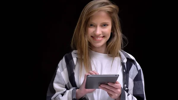 Portrait of young caucasian long-haired girl in shirt working with tablet and smiling into camera on black background. — Stock Photo, Image