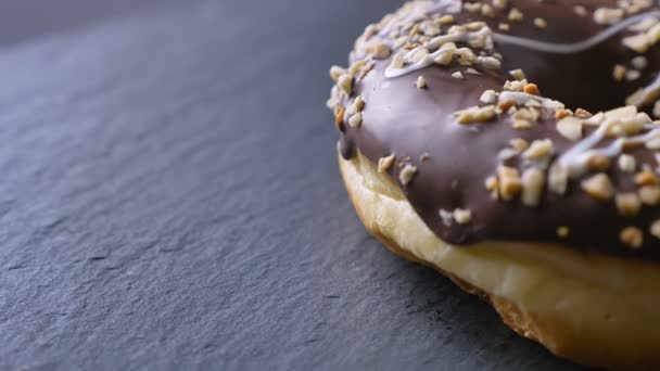 Close-up half-shot of glazed brown donut with crushed nuts spinning slowly on gray table background. — Stock Video