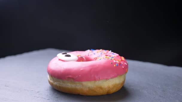 Close-up circulando em torno de tiro de delicioso donut rosa envidraçado com chips multicoloridos e olhos girando lentamente sobre fundo mesa cinza . — Vídeo de Stock