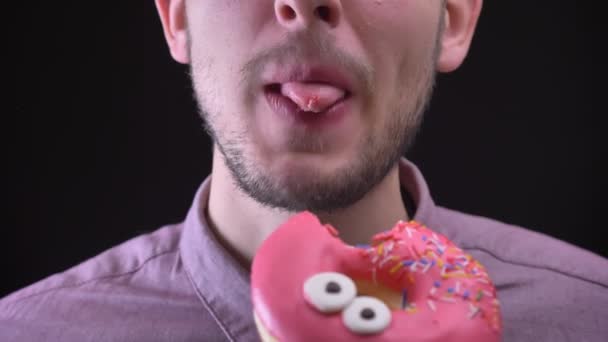 Close-up retrato de homem mordendo e mastigando delicioso donut rosa vitrificado com chips multicoloridos e olhos no fundo preto . — Vídeo de Stock