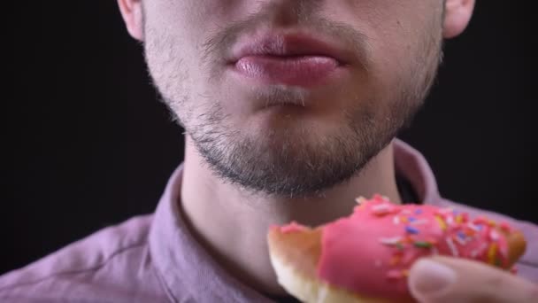 Close-up πορτρέτο του ανθρώπου το δάγκωμα και το μάσημα νόστιμα τζάμια ροζ ντόνατ με χρωματιστές μάρκες και τα μάτια σε μαύρο φόντο. — Αρχείο Βίντεο