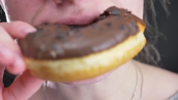 Close-up portrait of girl smilingly biting and chewing delicious glazed brown donut with chocolate chips on black background. — Stock Video