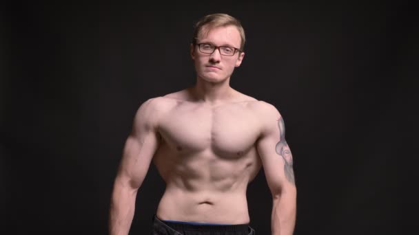 Portrait of naked young muscular man in glasses with tattoo watching calmly into camera on black background — Stock Video