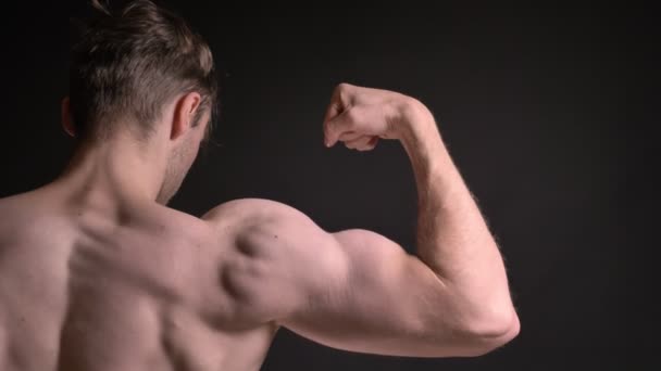 Retrato trasero de cerca del joven caucásico mostrando sus bíceps musculares y demostrando lo fuerte que es sobre fondo negro . — Vídeos de Stock