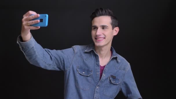 Closeup portrait of young caucasian man taking selfies on the phone with the background isolated on black — Stock Video