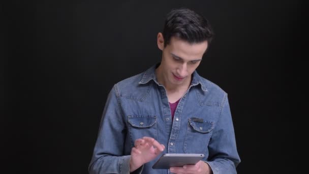Närbild porträtt av unga kaukasiska man att skriva på tabletten är distraherad och titta på kameran med bakgrund som isolerade på svart — Stockvideo