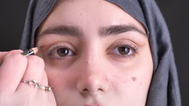 Close-up portret van vrouwelijke handen doen oog make-up met bruin potlood en penseel voor jonge moslimvrouw in hijab op zwarte achtergrond. — Stockvideo