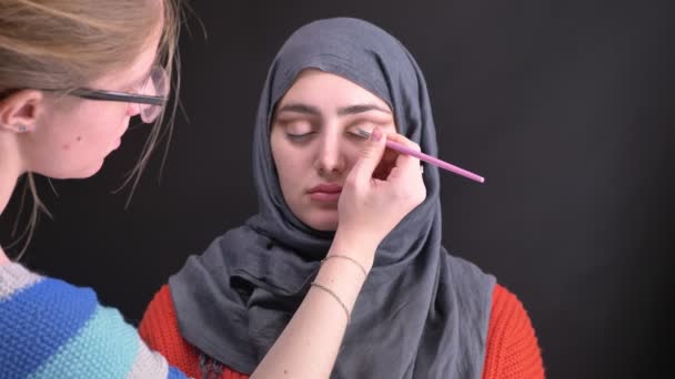 Retrato de manos femeninas haciendo maquillaje de ojos con cepillo pequeño para hermosa mujer musulmana en hijab sobre fondo negro . — Vídeo de stock