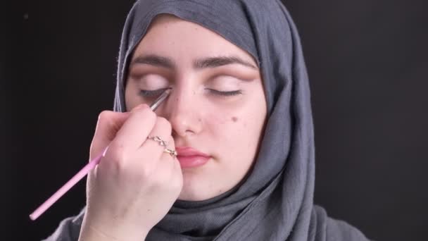 Retrato de manos femeninas maquillándose los ojos con un cepillo delgado para una hermosa mujer musulmana en hijab sobre fondo negro . — Vídeo de stock