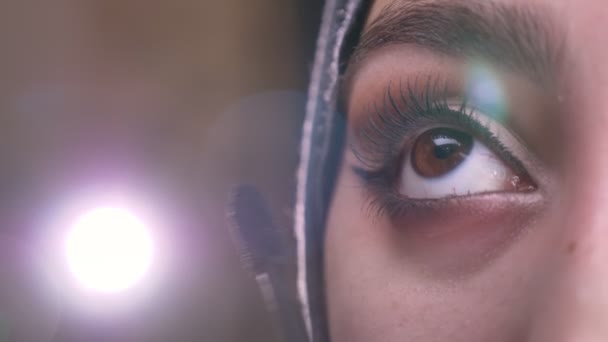 Gros plan portrait de mains féminines faisant maquillage à l'aide de mascara pour belle femme musulmane en hijab sur fond de lumières vives . — Video