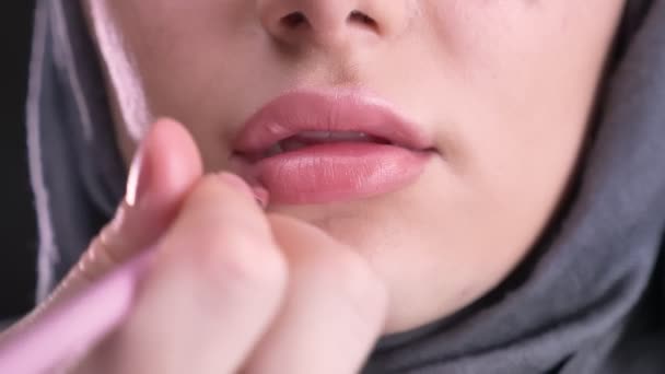 Närbild skott av processen av make-up och sätta rosa läppstift med tunn pensel på ung muslimsk tjej i hijab på svart bakgrund. — Stockvideo