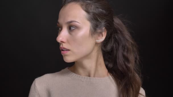 Close-up portret van jonge brunette Kaukasische meisje met paardenstaart draait zich naar de camera en horloges serieus erin op zwarte achtergrond. — Stockvideo