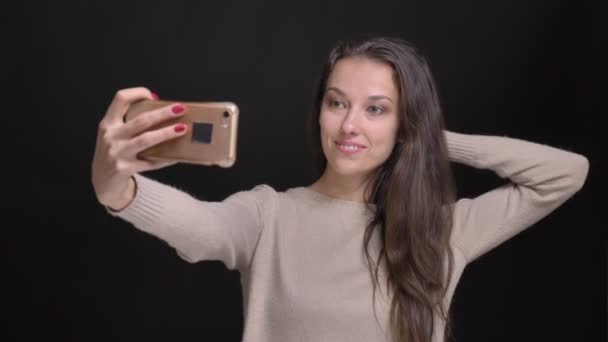 Portrait of young brunette long-haired caucasian girl joyfully making selfie-photos on smartphone on black background. — Stock Video