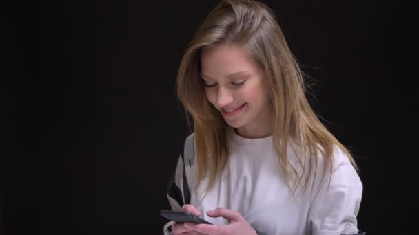 Retrato en el perfil de la joven caucásica de pelo largo chica en blanco camiseta relojes sonriendo en el teléfono inteligente sobre fondo negro . — Vídeo de stock