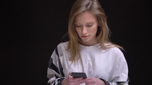 Retrato en el perfil de la joven caucásica de pelo largo chica en blanco camiseta relojes con la risa en el teléfono inteligente sobre fondo negro . — Vídeos de Stock