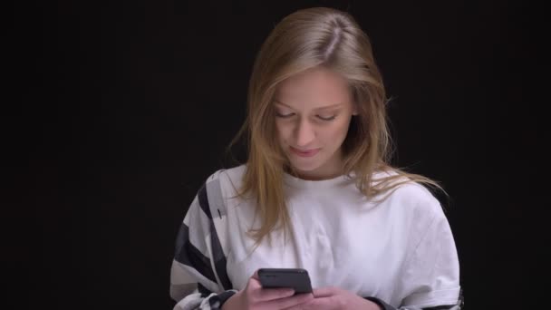 Retrato en el perfil de la rubia caucásica de pelo largo chica en blanco camiseta relojes con la risa en el teléfono inteligente sobre fondo negro . — Vídeos de Stock