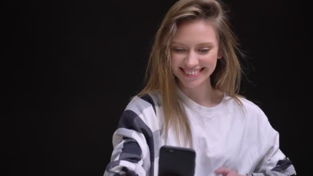 Retrato de rubia caucásica de pelo largo chica en camiseta blanca felizmente hablando en videochat en el teléfono inteligente sobre fondo negro . — Vídeo de stock