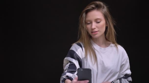 Retrato de rubia caucásica de pelo largo chica en camiseta blanca con mucho gusto hablando en videochat en el teléfono inteligente sobre fondo negro . — Vídeo de stock