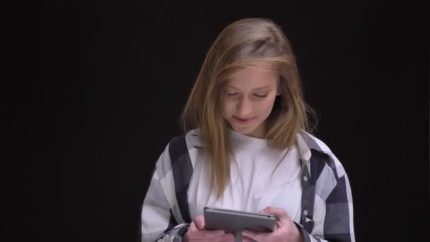 Blonde caucasian long-haired girl in plaid shirt smilingly working with tablet and joyfully watching into camera on black background. — Stock Video