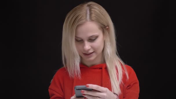 Retrato de una joven caucásica de pelo blanco en suéter rojo mirando sonriente en un smartphone sobre fondo negro . — Vídeos de Stock