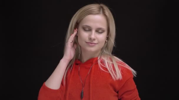Porträtt av ung vithårig kaukasiska flicka i röd tröja lyssnar på musik i hörlurar och sjunger på svart bakgrund. — Stockvideo