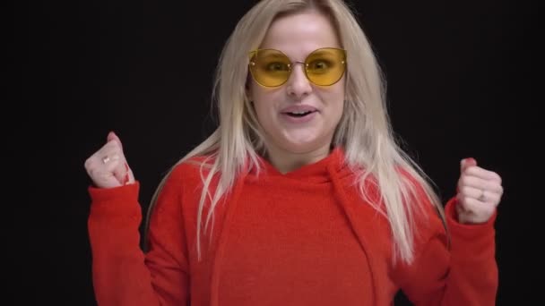 Portrait of young white-haired caucasian girl in yellow sunglasses gets extremely happy and jumps on black background. — Stock Video