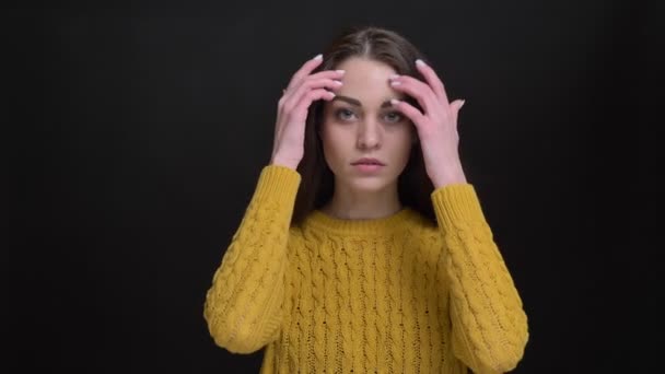 Portret van langharige brunette meisje in gele trui nederig tot vaststelling van haar haren en rustig kijken in de camera op zwarte achtergrond. — Stockvideo