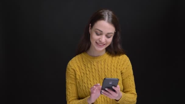 Retrato de chica morena de pelo largo en suéter amarillo trabajando alegremente con smartphone sobre fondo negro . — Vídeos de Stock