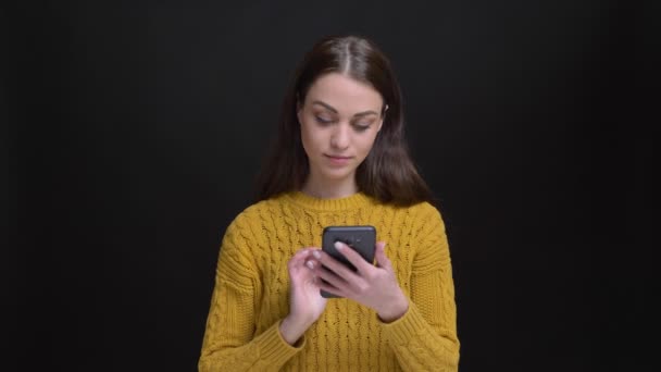 Длинноволосая брюнетка в желтом свитере улыбается, работает со смартфоном и смотрит в камеру на черном фоне . — стоковое видео