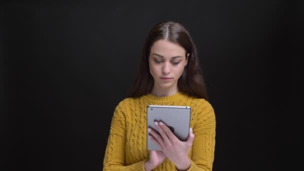 Retrato de menina morena de cabelos longos em suéter amarelo trabalhando atentamente com tablet e sorrindo para a câmera no fundo preto . — Vídeo de Stock