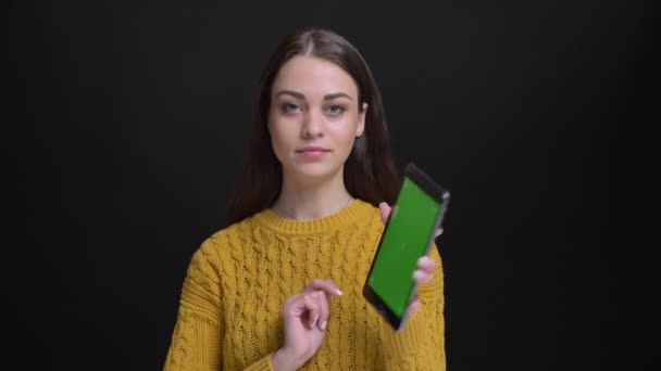Portret van langharige brunette meisje in gele trui vreugdevol tonen groen scherm van tablet in camera op zwarte achtergrond. — Stockvideo