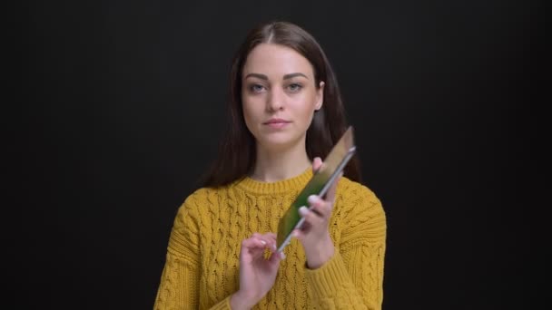 Portrait of long-haired brunette girl in yellow sweater smilingly showing green screen of tablet into camera on black background. — Stock Video