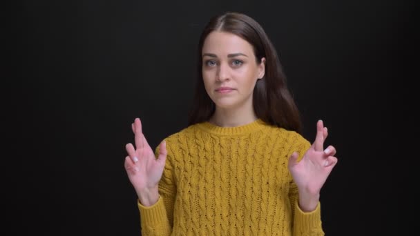 Retrato de menina morena de cabelos longos em suéter amarelo gesticulando sinal de dedos cruzados para mostrar esperança no fundo preto . — Vídeo de Stock