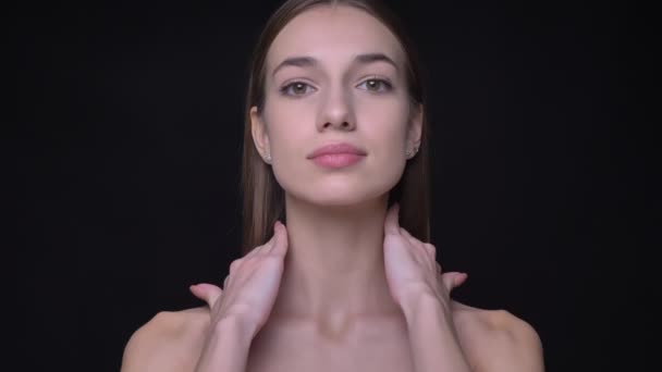 Portrait of young and slim caucasian girl with nude make-up watching into camera touching her collarbone on black background. — Stock Video