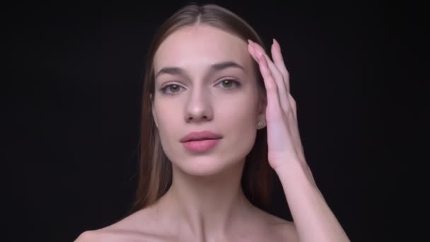 Portrait of young and slim caucasian girl with nude make-up watching into camera touching her face on black background. — Stock Video