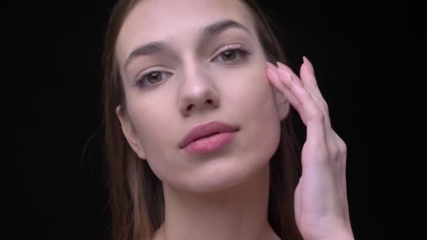 Close-up portrait of young caucasian girl with nude make-up watching into camera gently touching her face on black background. — Stock Video