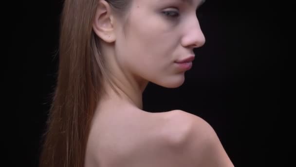 Porträt eines jungen und schlanken kaukasischen Mädchens mit nacktem Make-up wendet sich der Kamera zu und schaut ihr über die Schulter auf schwarzem Hintergrund zu. — Stockvideo