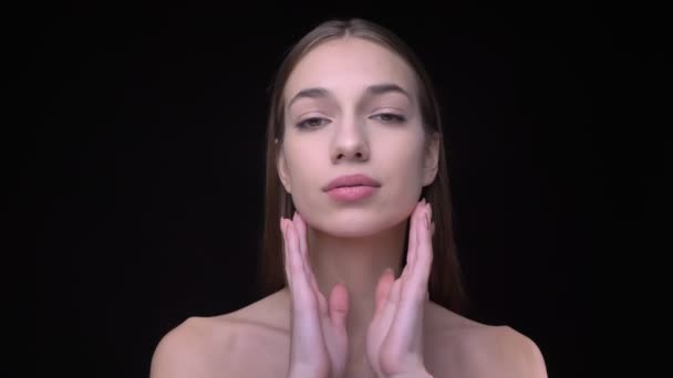 Young en slim Kaukasische meisje met naakt make-up kijken in de camera en het aanraken van haar gezicht en de kin op zwarte achtergrond. — Stockvideo