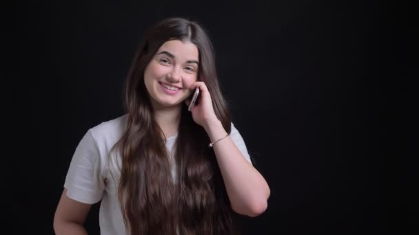 Portrait de jeune fille caucasienne brune en surpoids parlant joyeusement sur smartphone sur fond noir . — Video