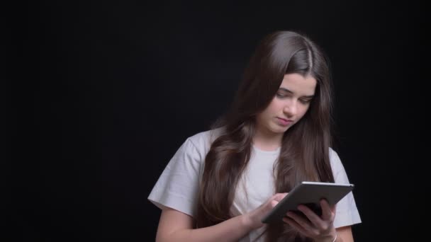 Portrait of young overweight brunette caucasian girl attentively working with tablet and smiling into camera on black background. — Stock Video