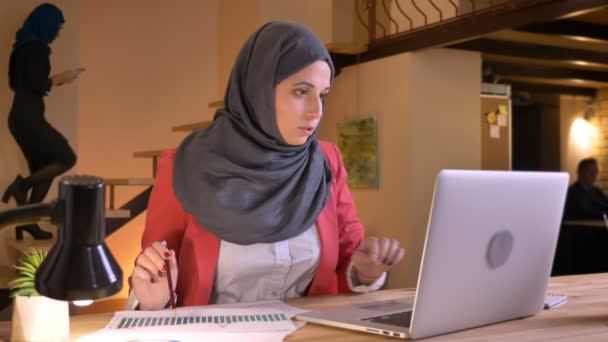 Крупним планом портрет молодої красивої мусульманської бізнес-леді в хіджабі, що працює в міжкультурній спільноті, використовуючи ноутбук, що вивчає графік і робить нотатки — стокове відео