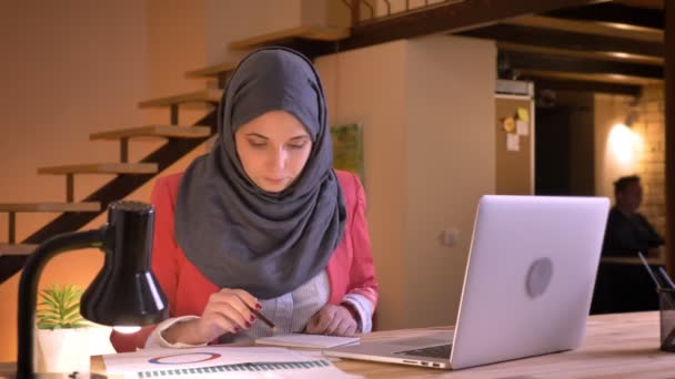 Närbild porträtt av unga uppmärksamma muslimska affärskvinna i hijab studerar data i diagrammet framför den bärbara datorn inomhus på arbetsplatsen — Stockvideo