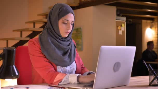 Retrato de close-up de jovem bela mulher de negócios muçulmana no hijab digitando no laptop olhando para a câmera e sorrindo alegremente dentro de casa no local de trabalho — Vídeo de Stock