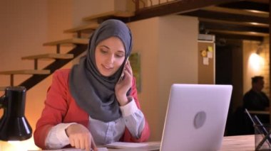 Genç başarılı Müslüman işkadını kapalı laptop kullanırken telefonda bir müşteri danışmanlık hijab içinde closeup portresi