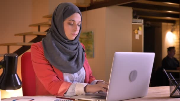 Vértes portréja hidzsáb gépelés-ra a laptop, kamera fordult, és mosolyogva, vidáman bírja a munkahelyen a fiatal gyönyörű muszlim üzletasszony — Stock videók