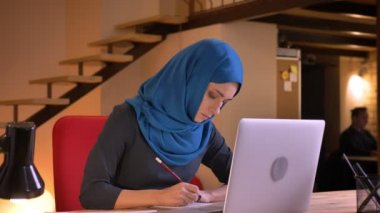 Genç ciddi Müslüman işkadını dizüstü ve kapalı çalışma alanı üzerinde not almak yazarak Türban içinde closeup portresi