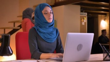Genç çekici Müslüman işkadını kameraya bakarak ve mutlu bir şekilde gülümseyerek dizüstü bilgisayarda yazarak closeup portresi