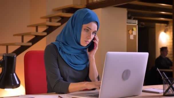 Retrato de cerca de una joven empleada árabe atractiva que tiene una conversación formal por teléfono mientras escribe en la computadora portátil — Vídeos de Stock